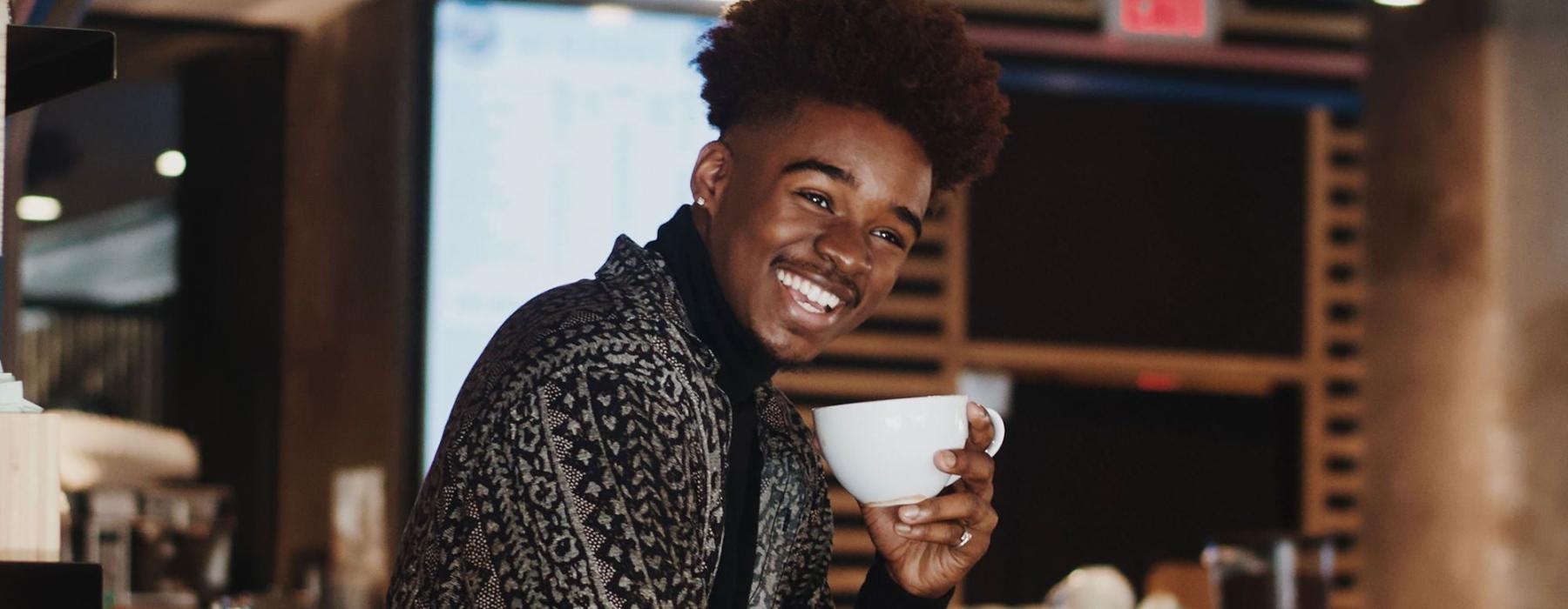 young man smiles while holding a cup of coffee in coffee shop